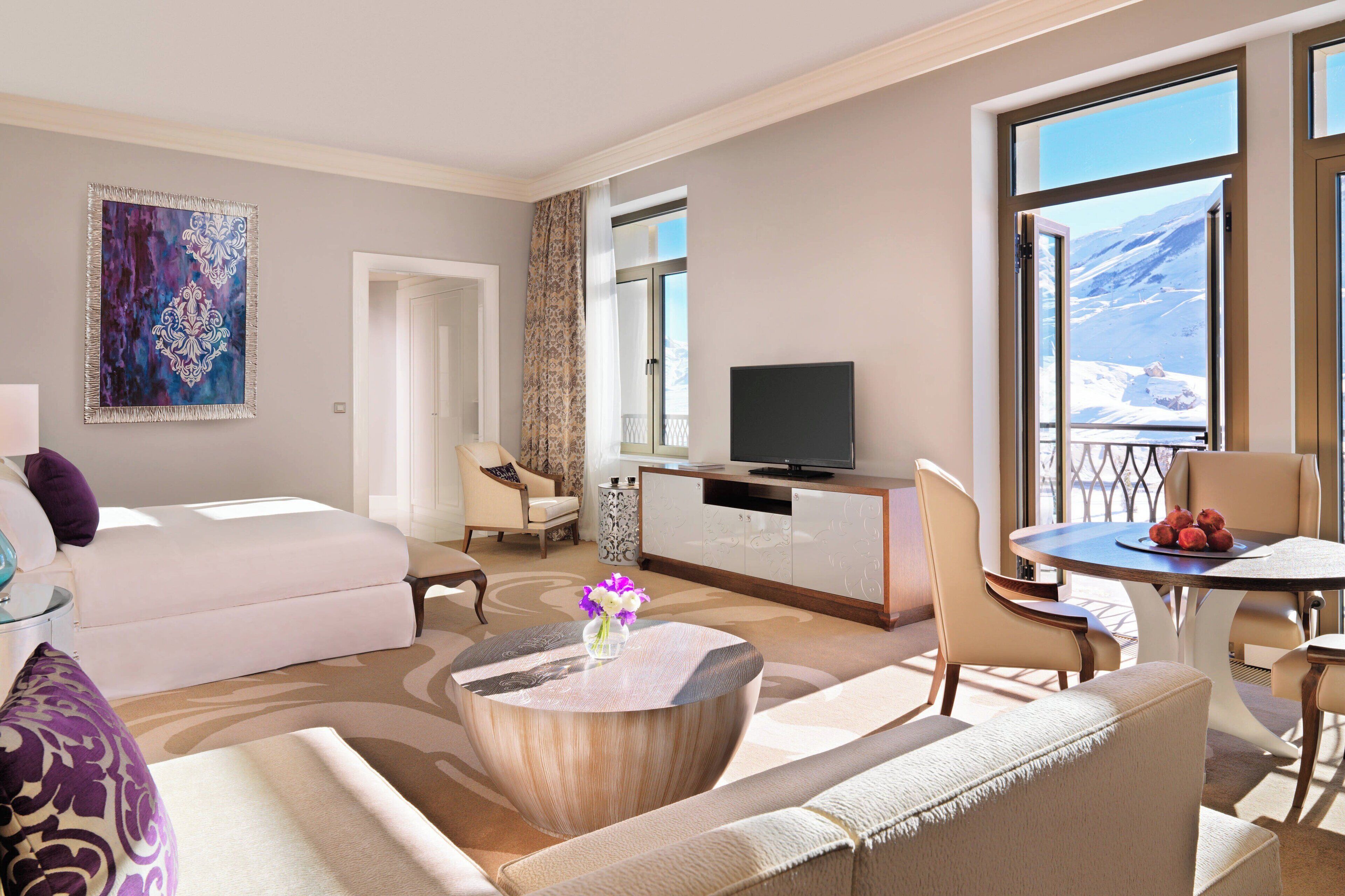 A modern hotel room with a bed, a sitting area with a coffee table, a TV, and a balcony with a mountain view.