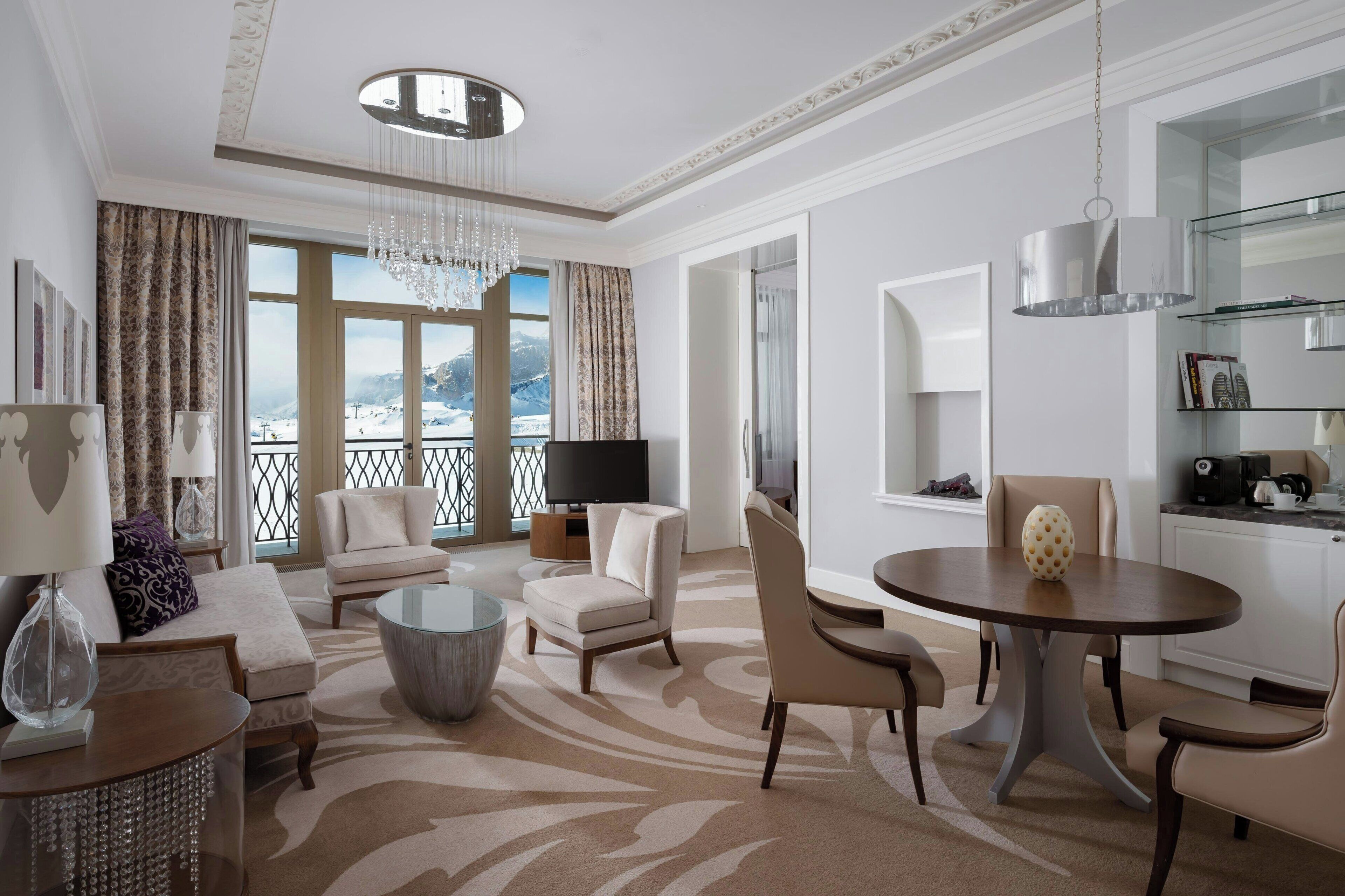A modern hotel room with a large window offering a mountain view, a chandelier, a flat-screen TV, and a dining area with a round table.