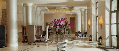 A spacious lobby with a grand chandelier, marble columns, and a central floral arrangement.