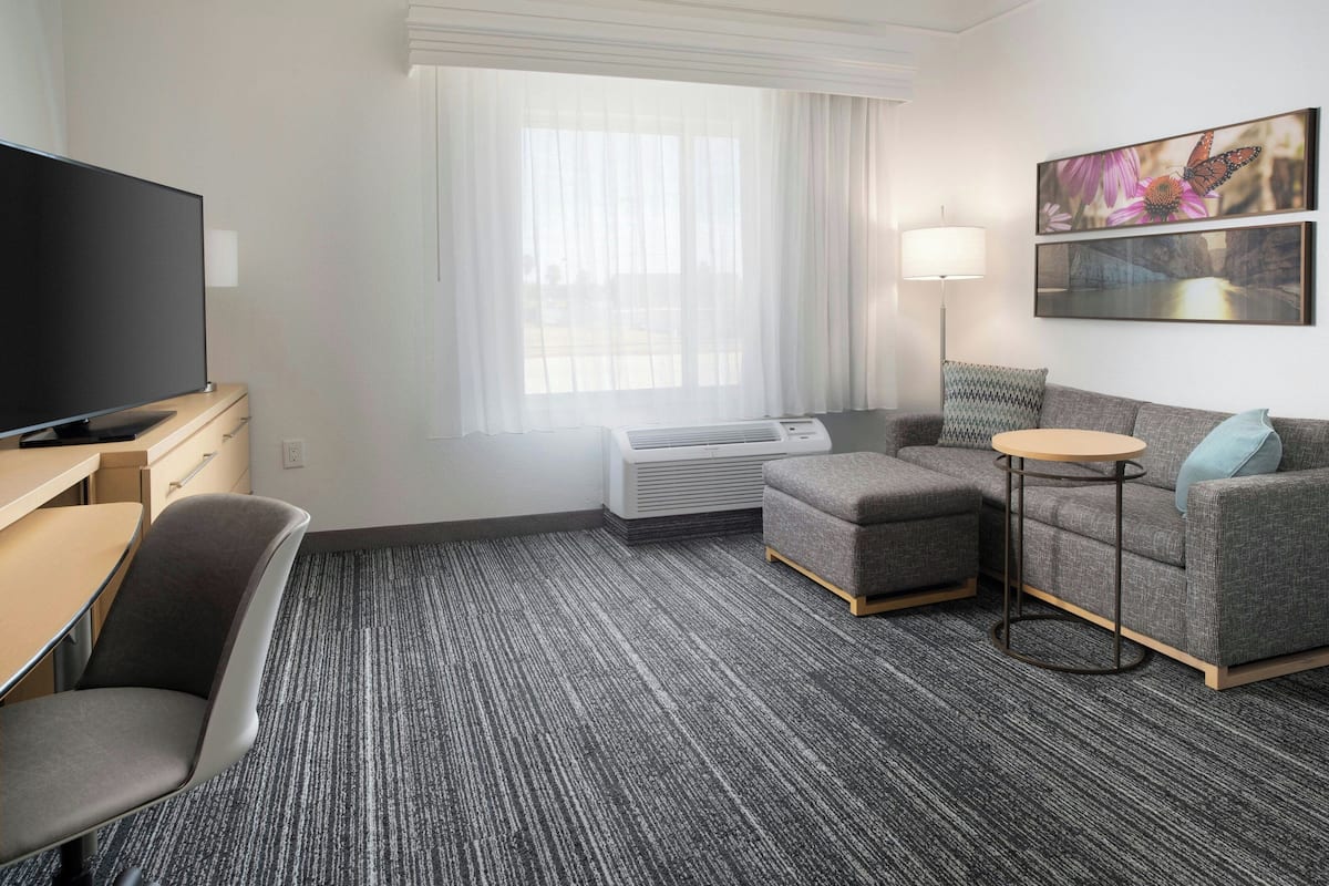 Habitación de hotel con televisión de pantalla plana, un escritorio, un sofá gris, una mesita redonda pequeña y un ventanal con cortinas translúcidas.