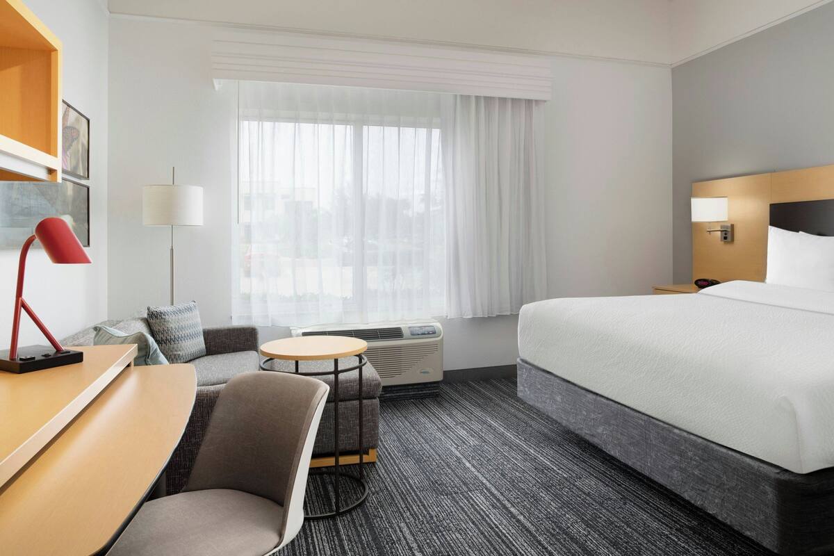 Habitación de hotel con cama, escritorio, silla, mesita, lámpara y ventana con cortinas.