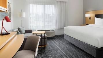 Habitación de hotel con cama, escritorio, silla, mesita, lámpara y ventana con cortinas.