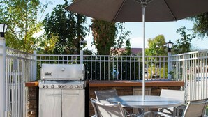 An outdoor patio with a grill, white metal chairs, and a table under a large umbrella. - TownePlace Suites by Marriott Laredo (Laredo)