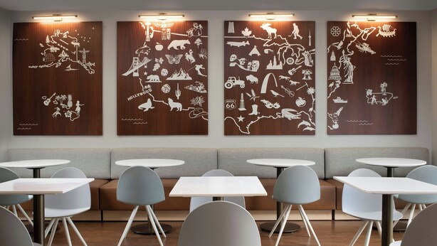 A modern dining area with round tables and grey chairs, a sofa, and wall art depicting various scenes. - TownePlace Suites by Marriott Laredo (Laredo)