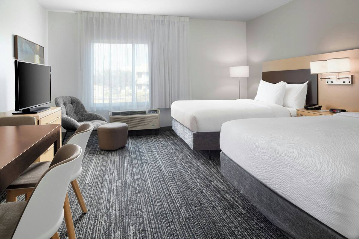 Habitación de hotel con dos camas, un escritorio con televisión, una silla, una lámpara y una ventana con cortinas.