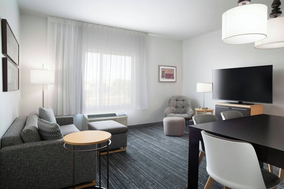 Habitación de hotel moderna con un sofá gris, una mesa redonda, un televisor de pantalla plana y un amplio ventanal con cortinas blancas.