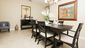A dining area with a dark wooden table, six chairs, a vase of white flowers, a chandelier, and a framed painting on the wall.