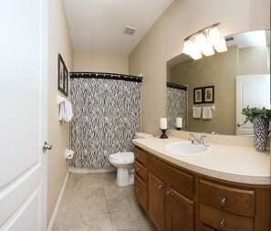 A bathroom with a zebra-striped shower curtain, a white sink, wooden cabinets, and a mirror with lights above it. - Highlands Reserve Homes (Davenport)