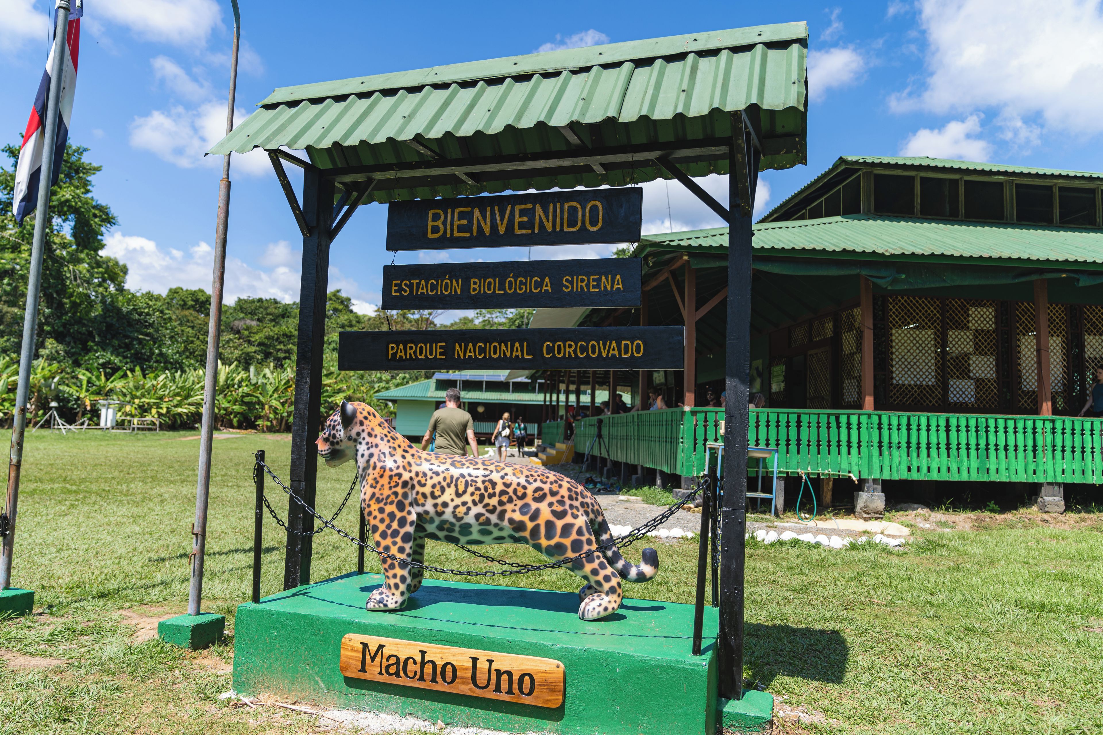 Un letrero de bienvenida con una estatua de jaguar, un edificio de techo verde y un cartel que dice 'Macho Uno'.