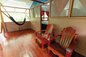 Ein Zimmer mit einer Hängematte, zwei Holzstühlen, einem Bett mit blauer Decke und einem Fenster mit roten Vorhängen.