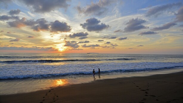 Cena de praia com pegadas que levam à água, um pôr do sol e duas pessoas à beira-mar.