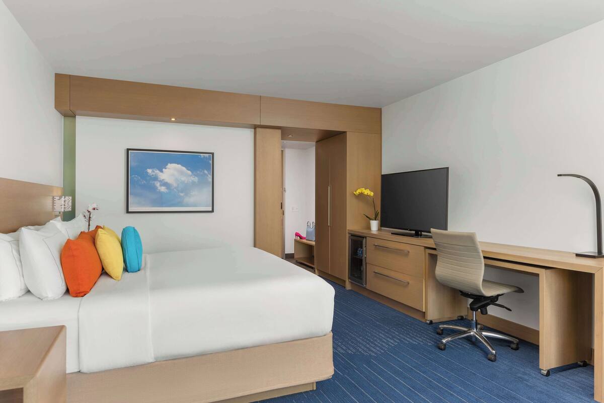 A hotel room with a bed, a desk, a television, and a window with a view of the sky.