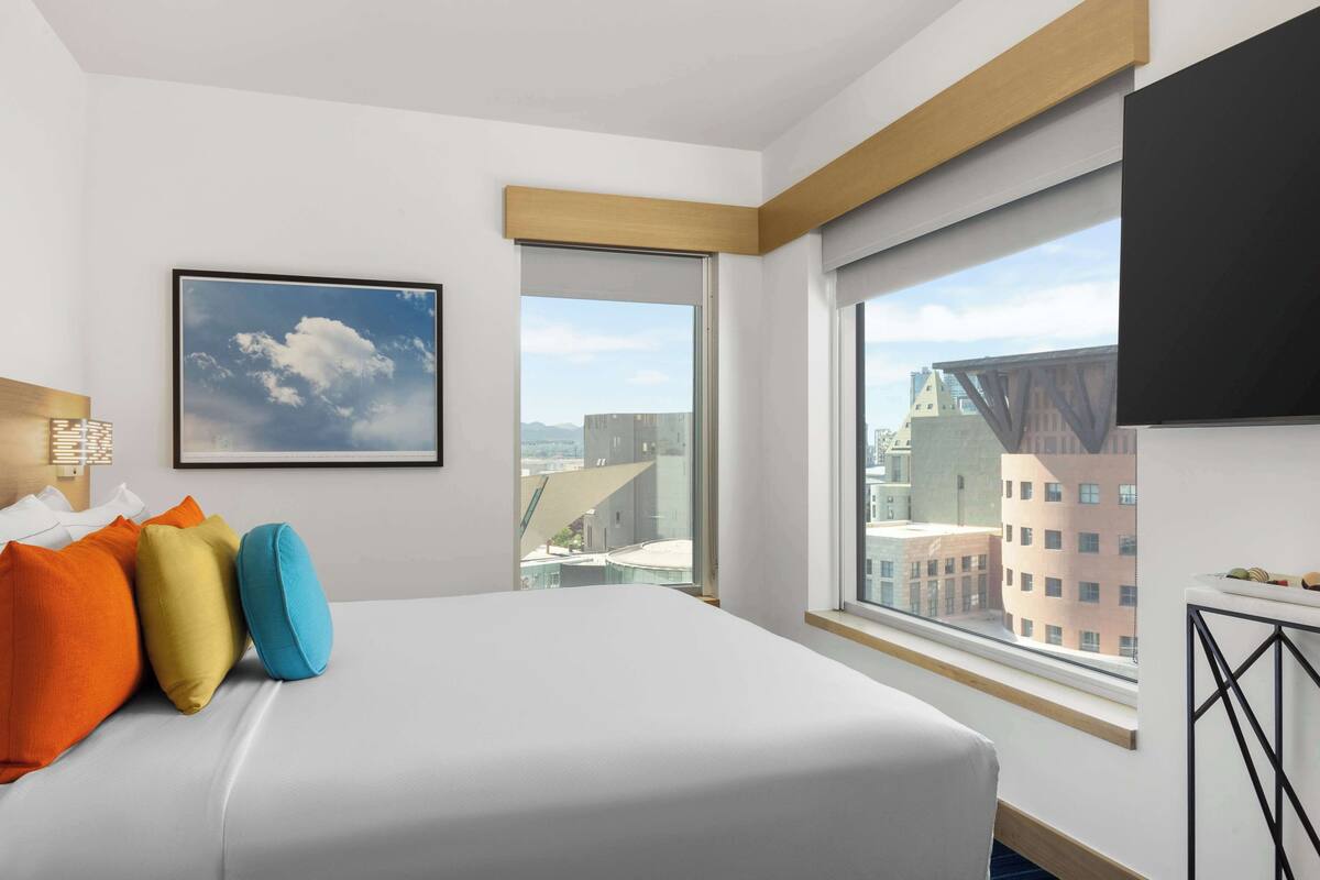 A hotel room with a large bed, a city view, a framed picture on the wall, and a window with a view of buildings.