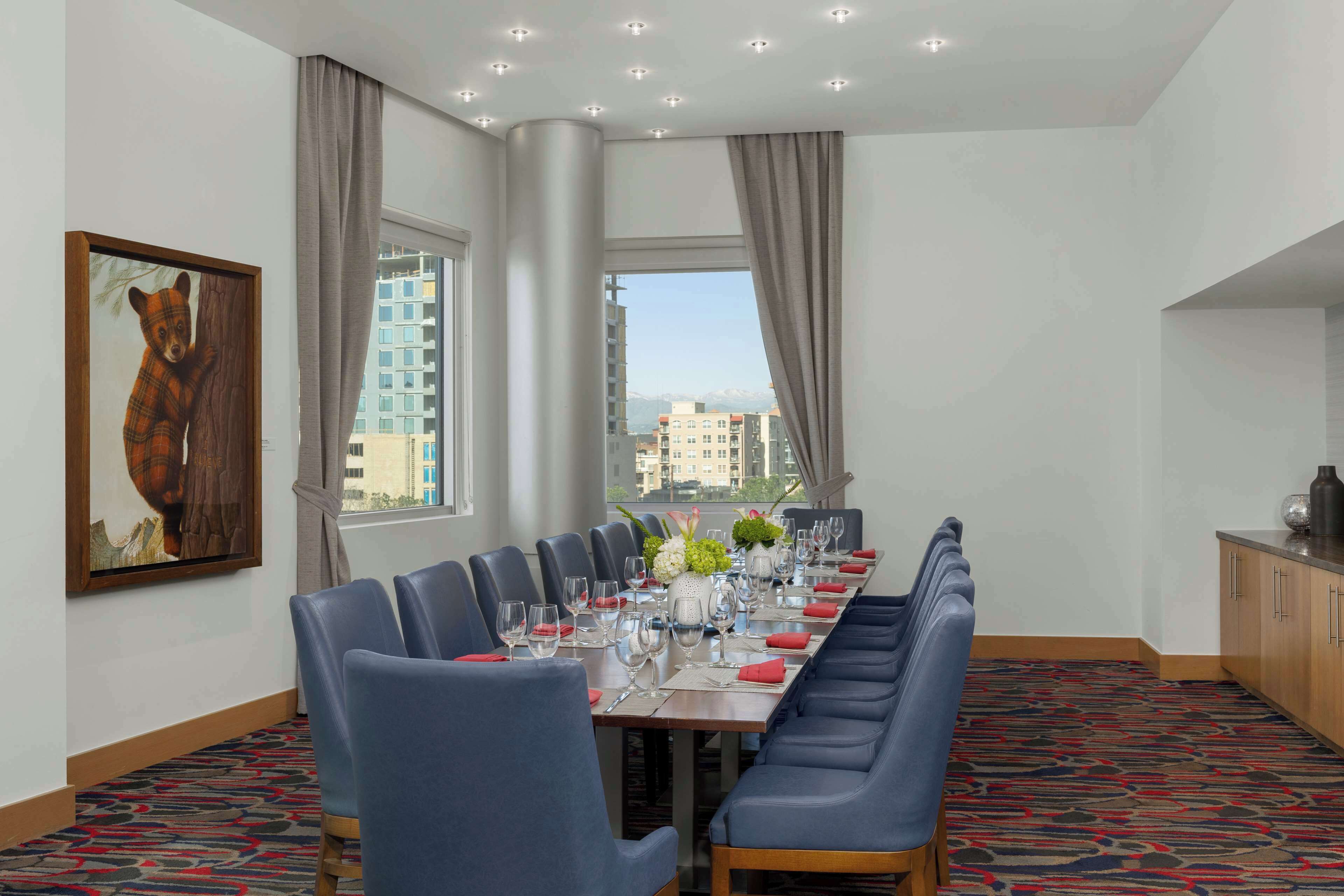 Un comedor de hotel con una larga mesa dispuesta para una comida, sillas azules, un cuadro en la pared y vistas de edificios a través de las ventanas.
