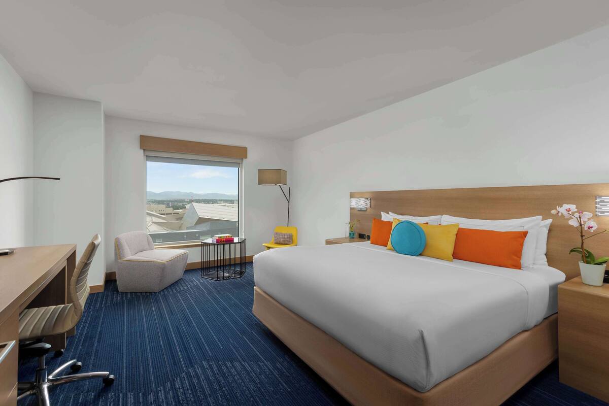 A hotel room with a large bed, a desk, a chair, a window with a view, and a nightstand with a lamp.