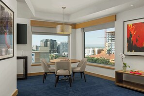 A modern hotel room with a round dining table, two chairs, a hanging light fixture, large windows with a city view, and a bookshelf with books and a vase of flowers. - The Art Hotel Denver, Curio Collection by Hilton (Denver)