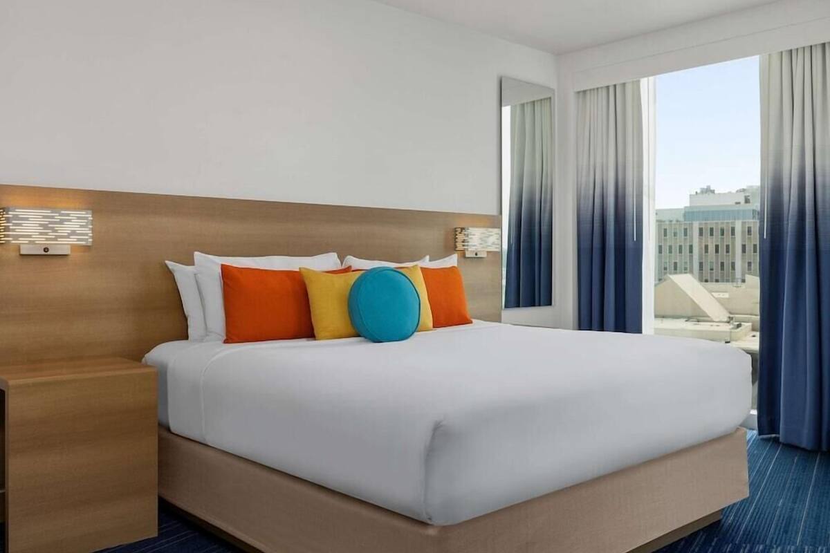 A hotel room with a large bed, a wooden headboard, a nightstand, and a view of buildings through the window.