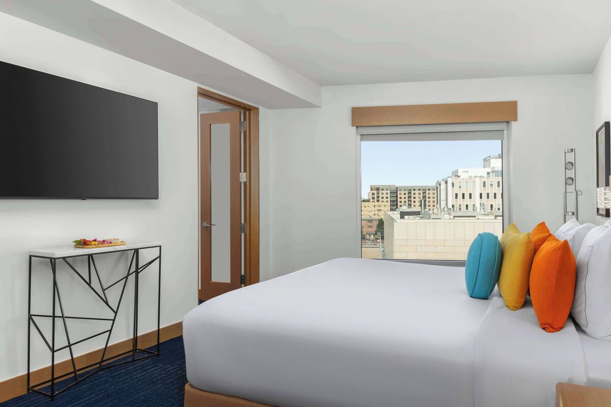 A hotel room with a large bed, a flat-screen TV mounted on the wall, a small side table, and a window with a city view.