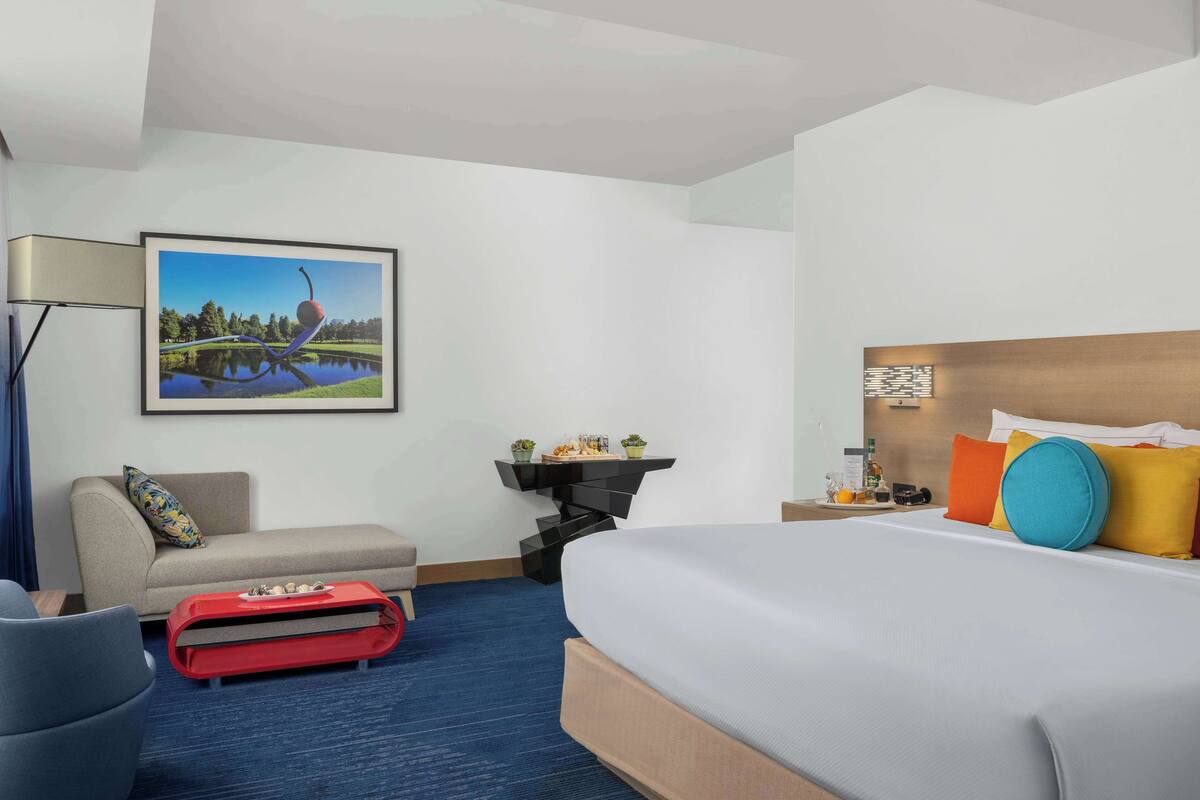 A hotel room with a bed, a sofa, a red luggage trolley, a desk with a lamp, and a framed picture on the wall.