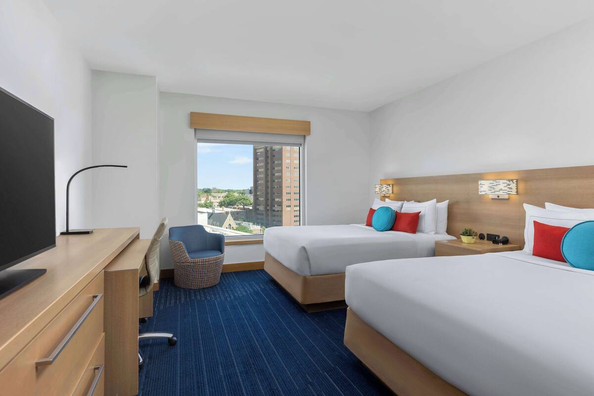 A hotel room with two beds, a desk, a TV, a chair, and a window with a city view.