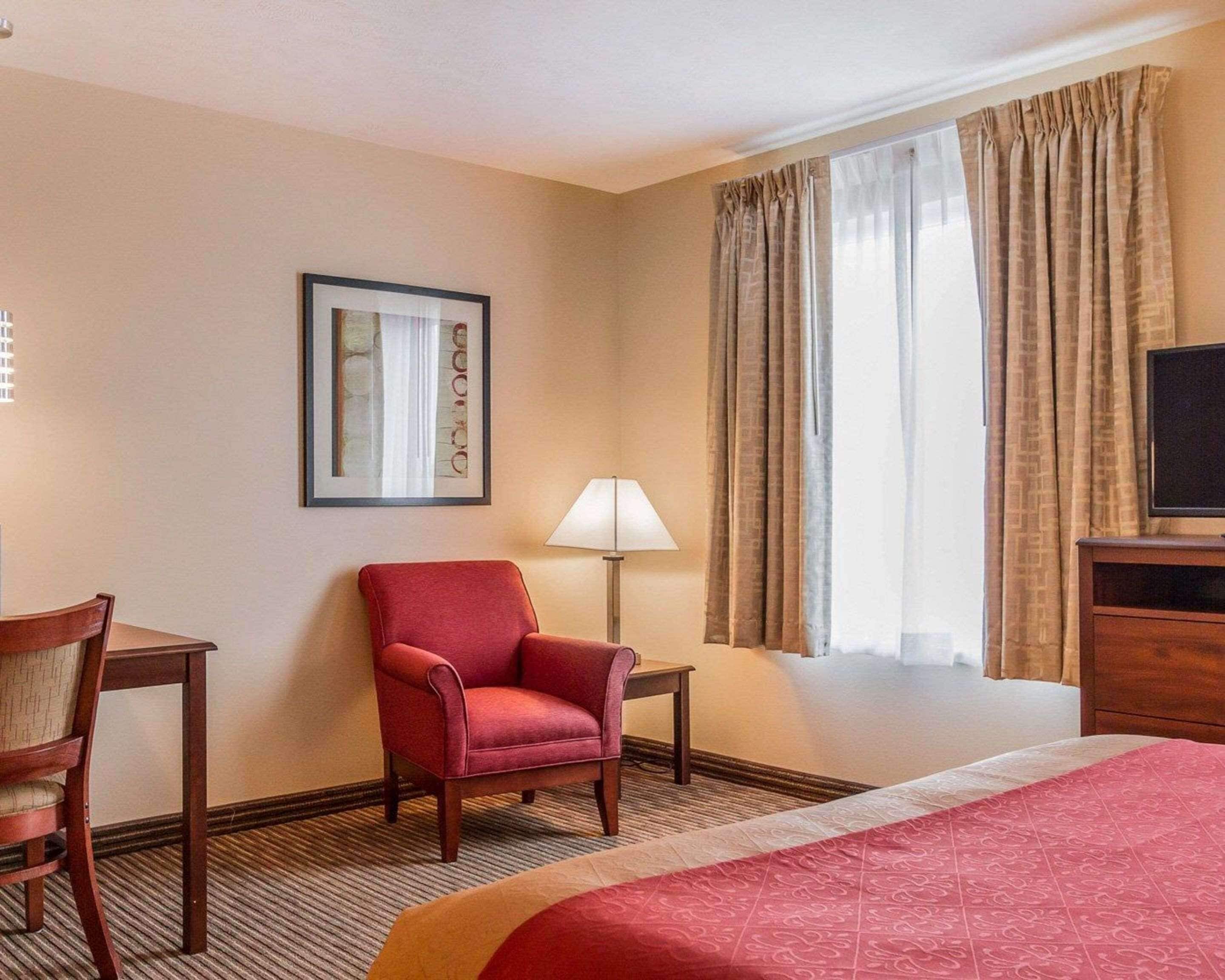 Una habitación de hotel con una cama, un sillón rojo, un escritorio con silla, una lámpara, un televisor y una ventana con cortinas.