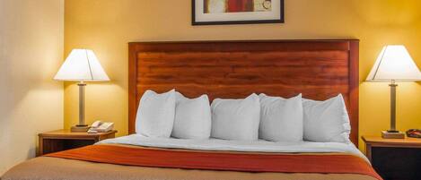 Habitación de hotel con cabecera de madera, cama con almohadas blancas, colcha marrón, dos mesitas de noche con lámparas y una obra de arte enmarcada en la pared.