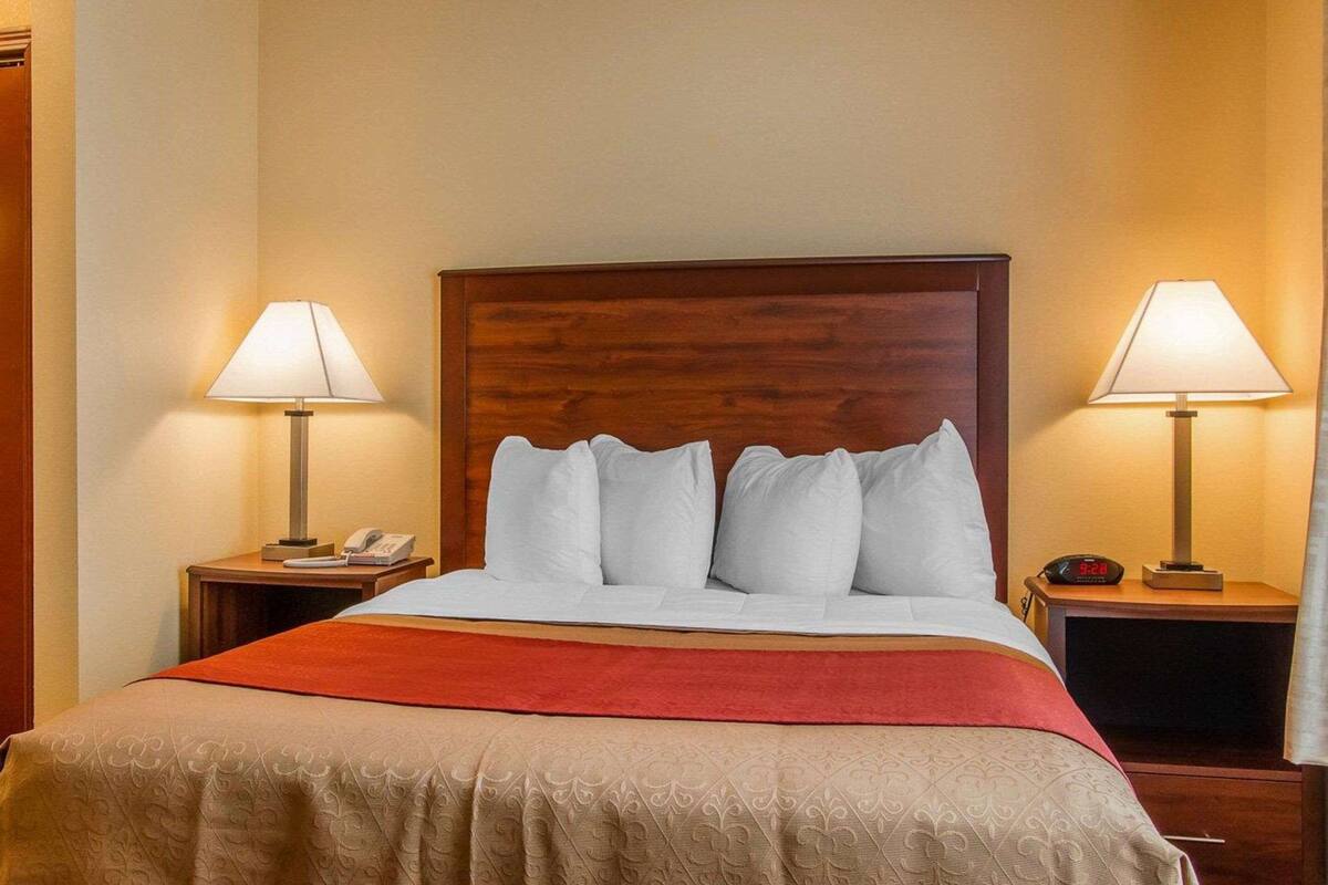 A hotel room with a large bed, two bedside tables with lamps, a red throw blanket, and a clock on one of the tables.