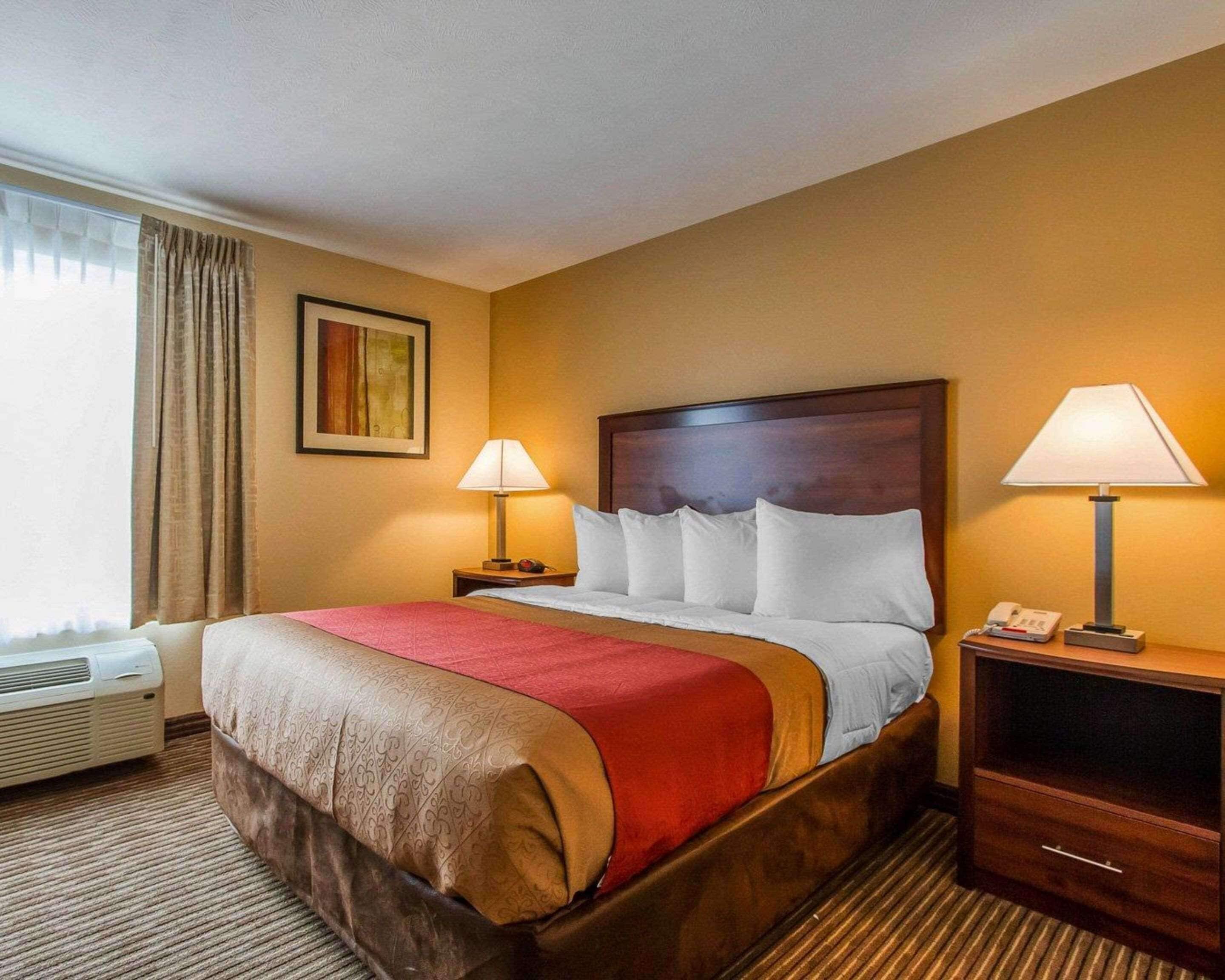 A hotel room with a large bed, two bedside tables, a heater, a window with curtains, and a painting on the wall.