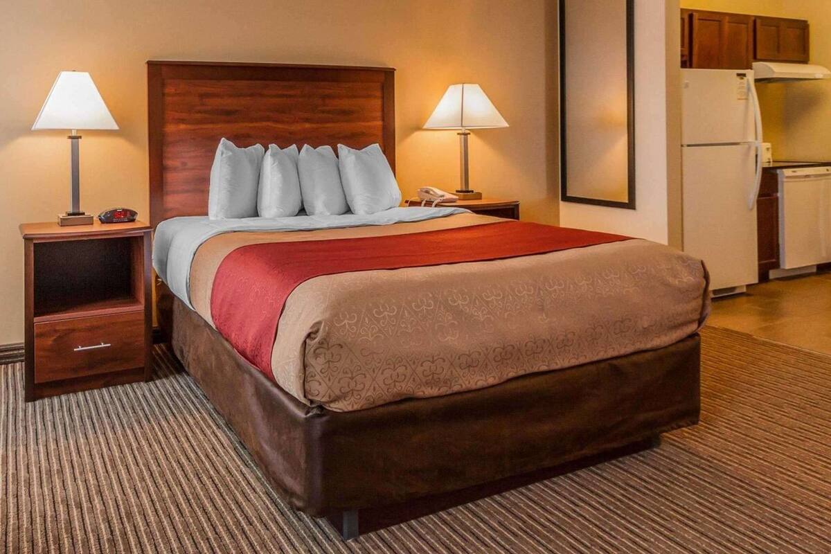 A hotel room with a bed, two bedside tables, a nightstand, a lamp, a phone, a red throw blanket, a wooden headboard, a framed picture on the wall, and a kitchenette with a refrigerator and stove.