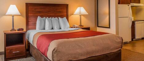 Habitación de hotel con cama, dos mesitas de noche, lámpara, teléfono, manta roja, cabecero de madera, un cuadro enmarcado en la pared y una kitchenette con refrigerador y estufa.
