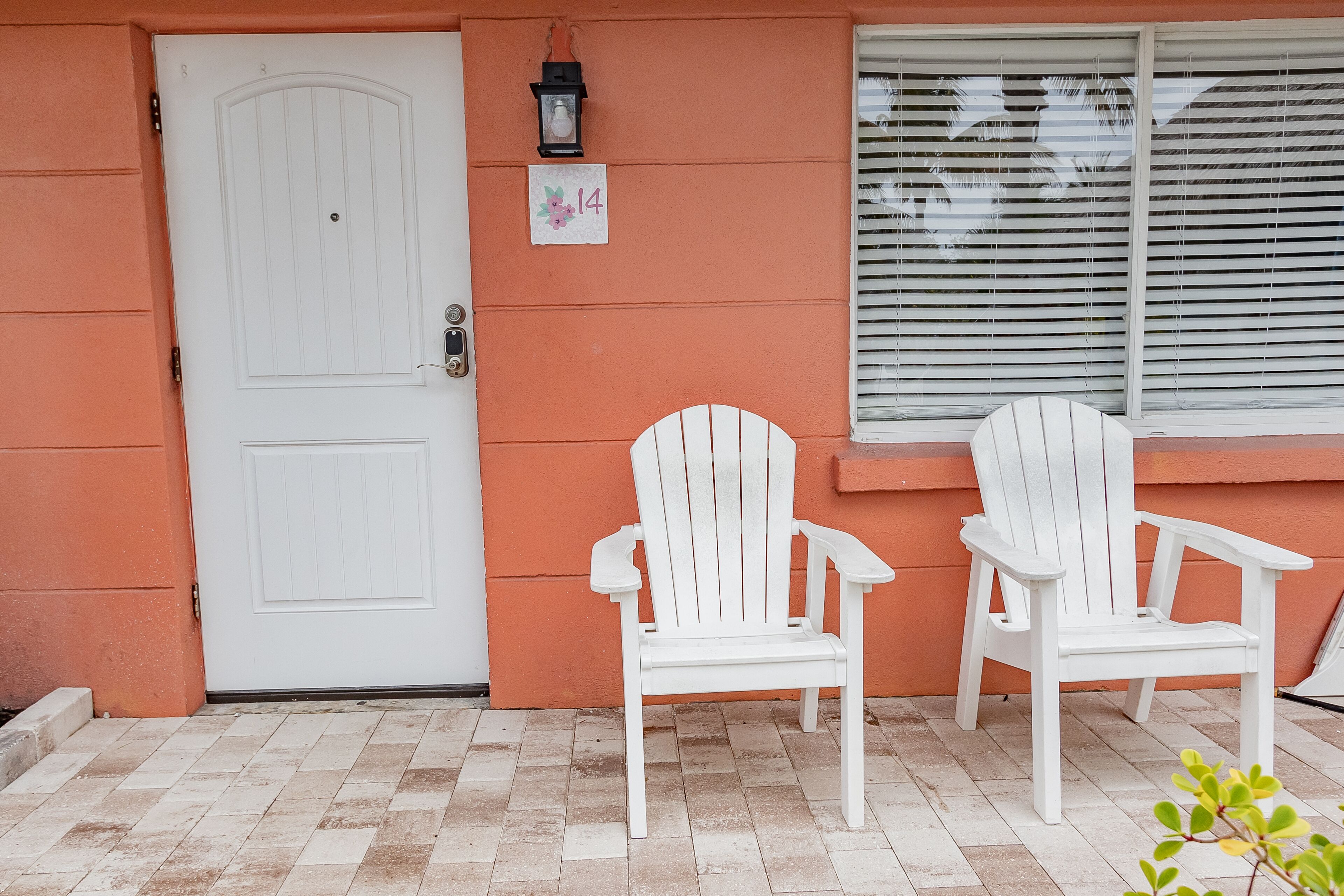 Une porte blanche, deux fauteuils Adirondack blancs et une fenêtre avec des stores.