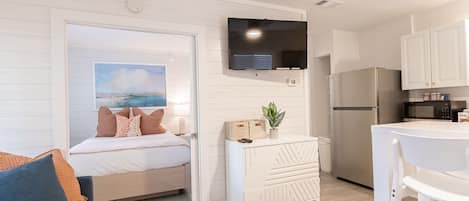 A modern room with a bed, a white dresser, a television, a kitchen area with a refrigerator and microwave, and a view of the ocean through a window.