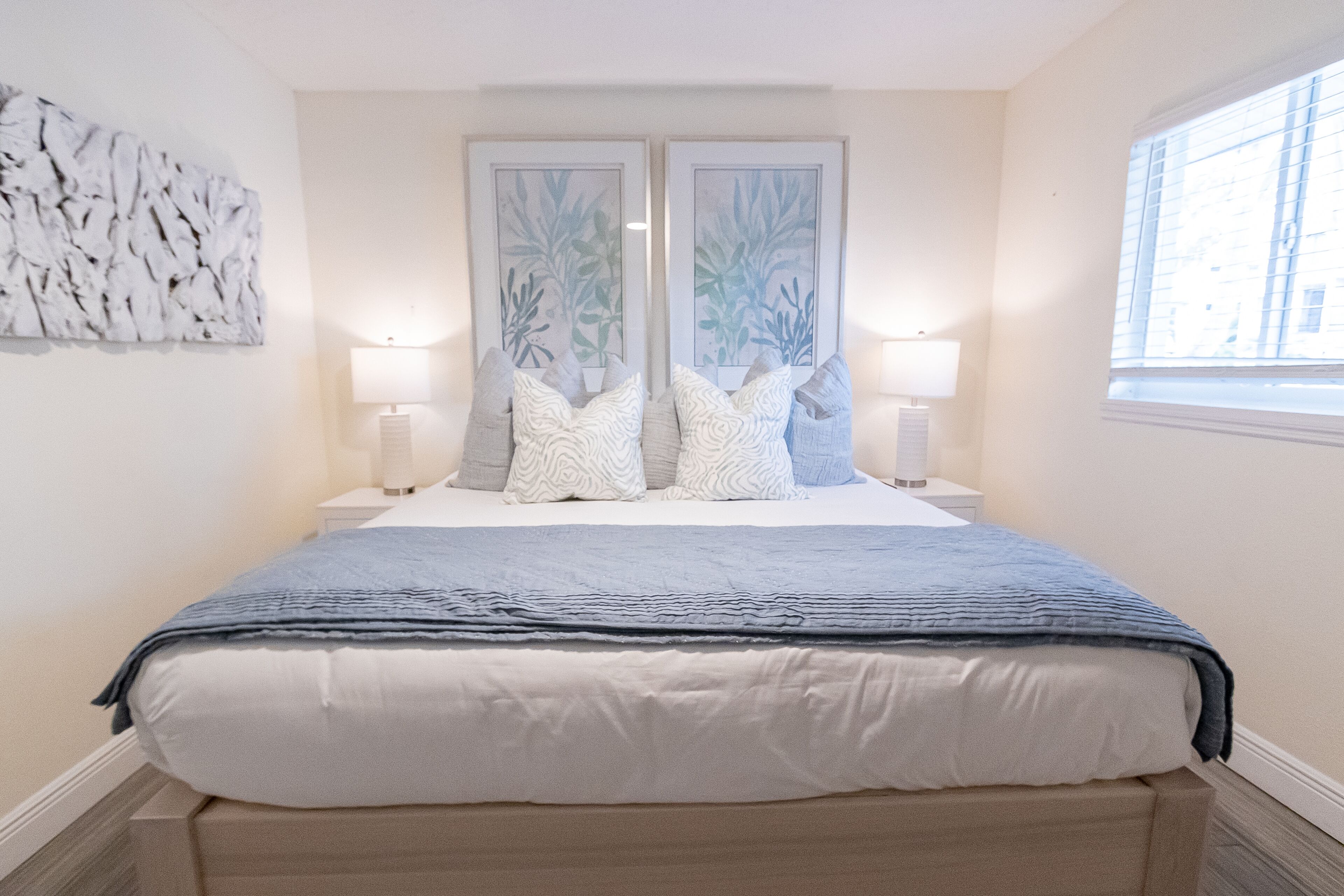 A neatly made bed with a blue blanket, two framed botanical prints above it, and a window on the right.