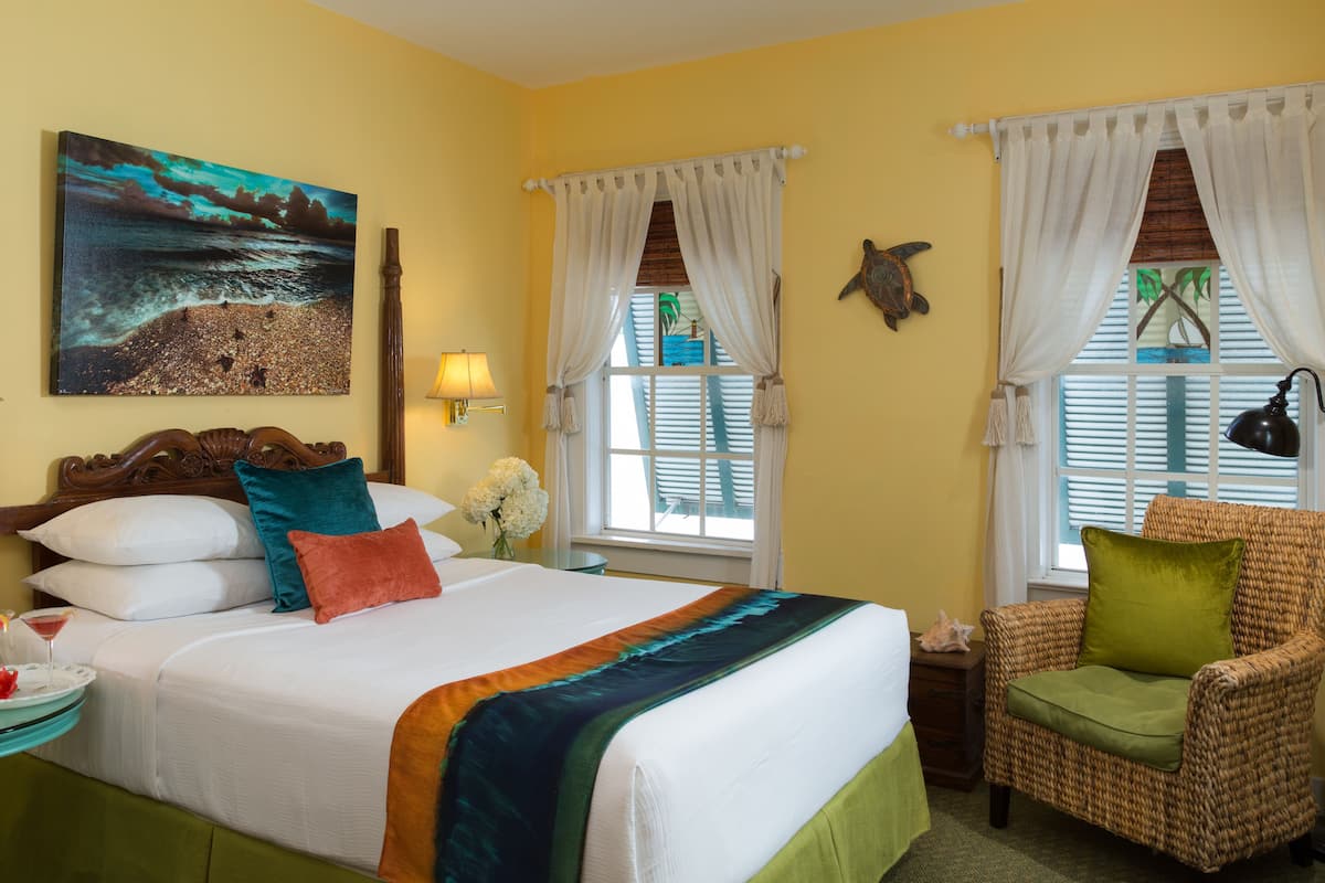 A hotel room with a bed, a chair, a painting, a lamp, and a window with curtains.