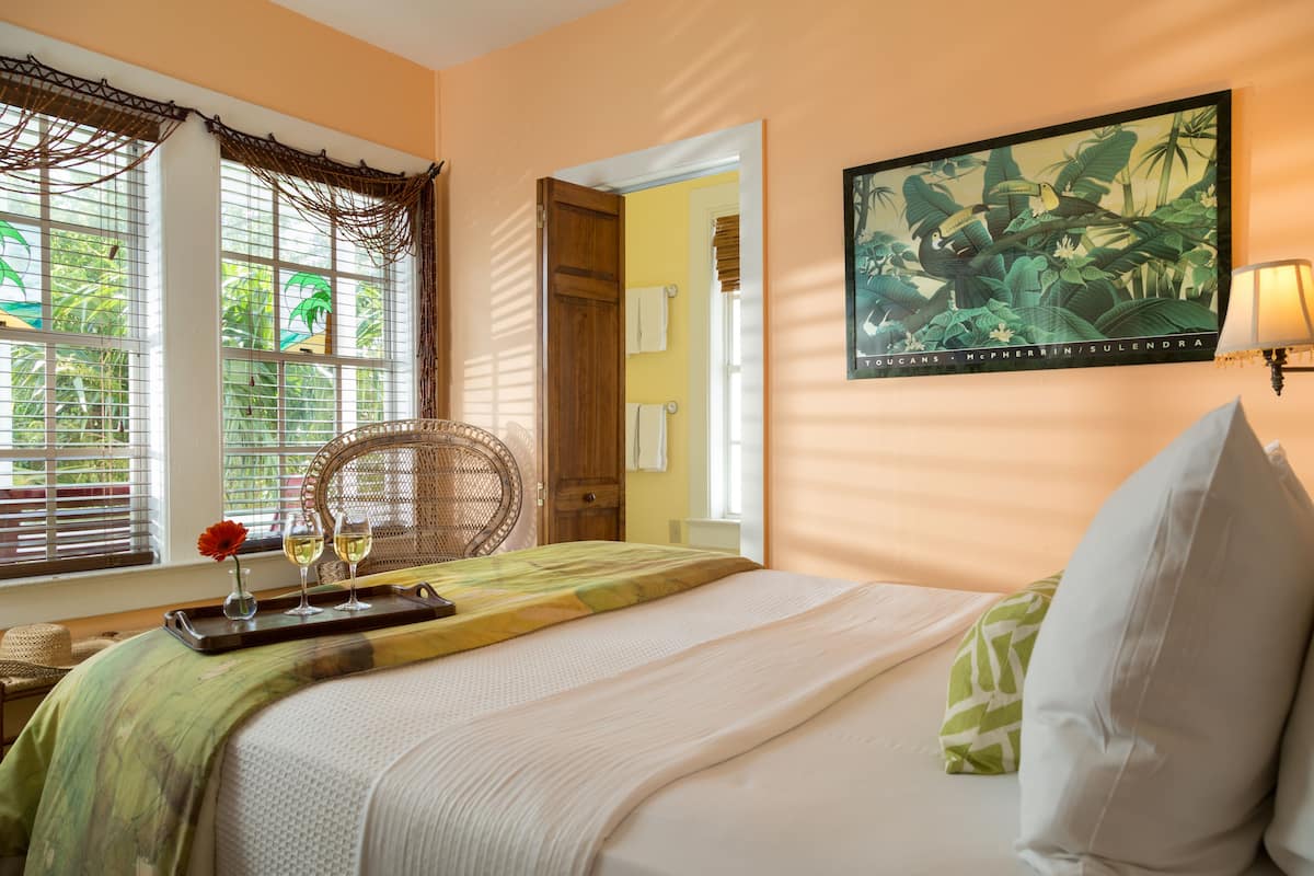 A hotel room with a bed, a chair, a tray with glasses and a flower, and a painting on the wall.