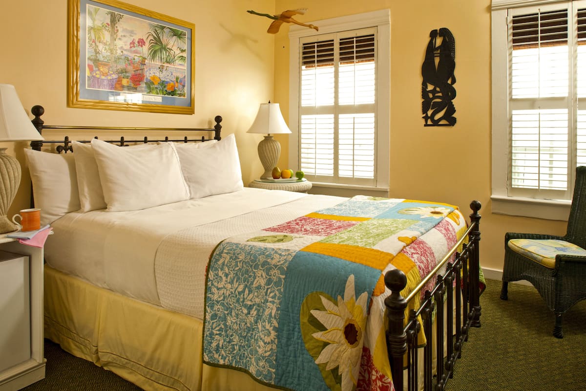 A neatly made bed with a floral quilt, a bedside table with a lamp, and a chair in a room with a window and a wall decoration.