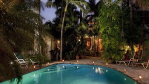 Un área de piscina con sillones de descanso y velas, rodeada de palmeras y un edificio al fondo.