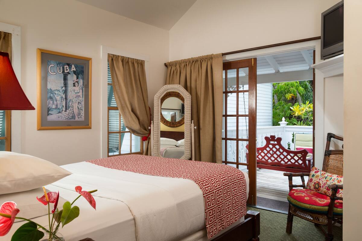 A hotel room with a bed, a mirror, a chair, and a door leading to a balcony with plants.