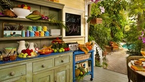 Un bufé de desayuno con una variedad de frutas, pasteles y bebidas en una terraza de madera con vistas a un jardín exuberante y una piscina.