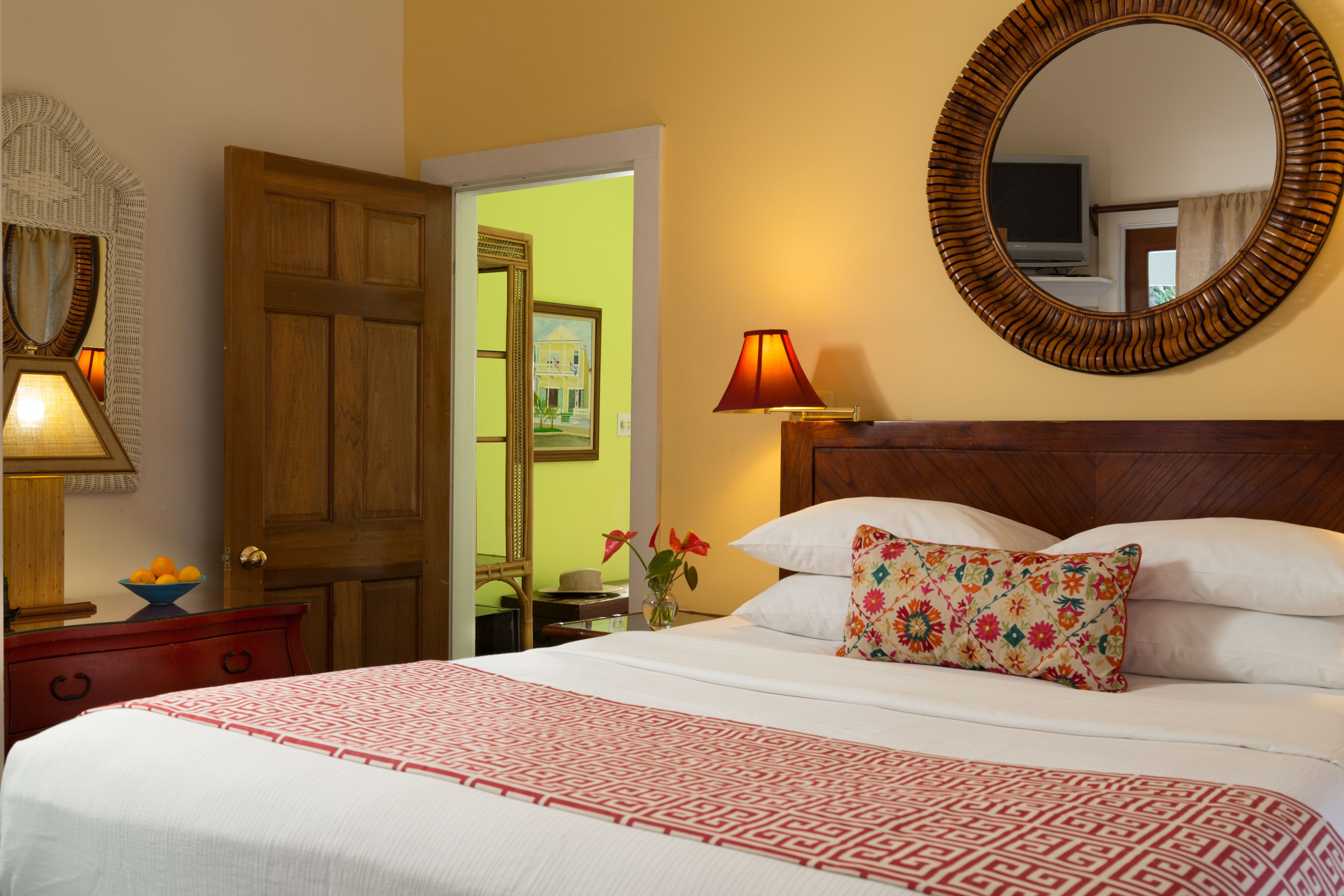 A neatly made bed with a patterned bedspread, a wooden headboard, and a floral pillow. There is a wooden nightstand with a lamp and a bowl of fruit. A mirror with a wooden frame is on the wall, and a door is visible in the background.
