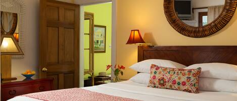 A neatly made bed with a patterned bedspread, a wooden headboard, and a floral pillow. There is a wooden nightstand with a lamp and a bowl of fruit. A mirror with a wooden frame is on the wall, and a door is visible in the background.