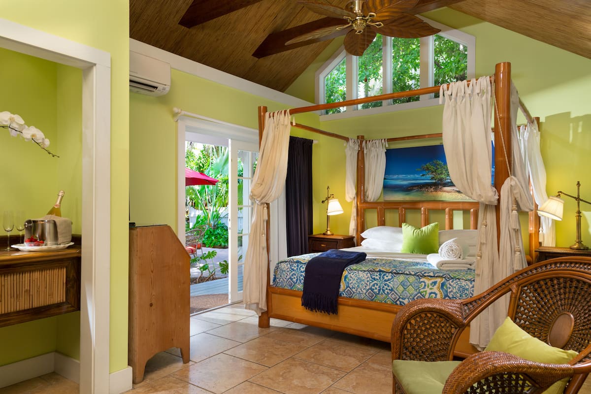 A bedroom with a four-poster bed, a wooden desk, wicker chairs, and a view of the outdoors.