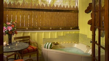 A bathroom with a bathtub, a window with a bamboo blind, a table with a vase of flowers, and a chair with striped cushions.
