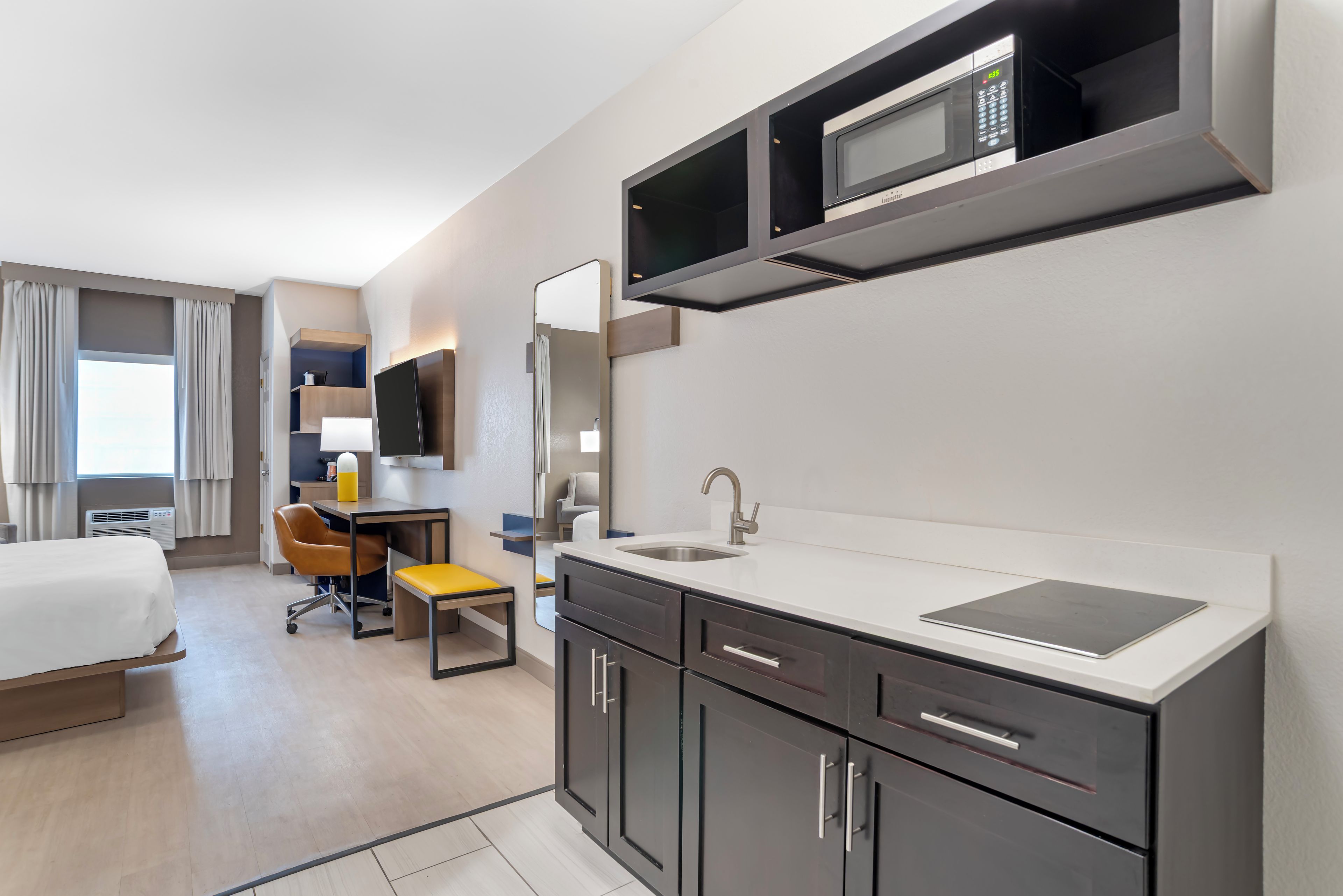 A modern hotel room with a bed, a desk, a chair, a bathroom, and a kitchenette.