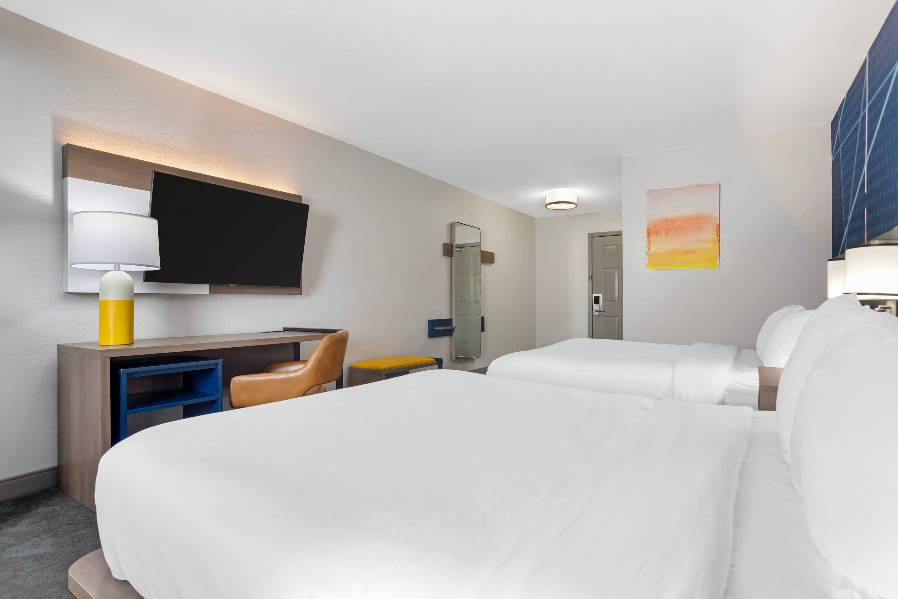 A hotel room with two beds, a desk, a chair, a TV, and a painting on the wall.