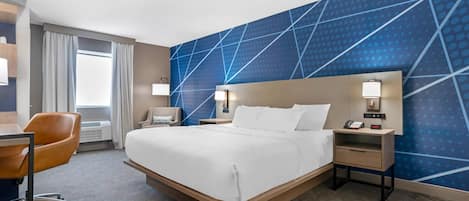 A hotel room with a large bed, a desk with a chair, a nightstand with a lamp, and a wall with a geometric pattern.
