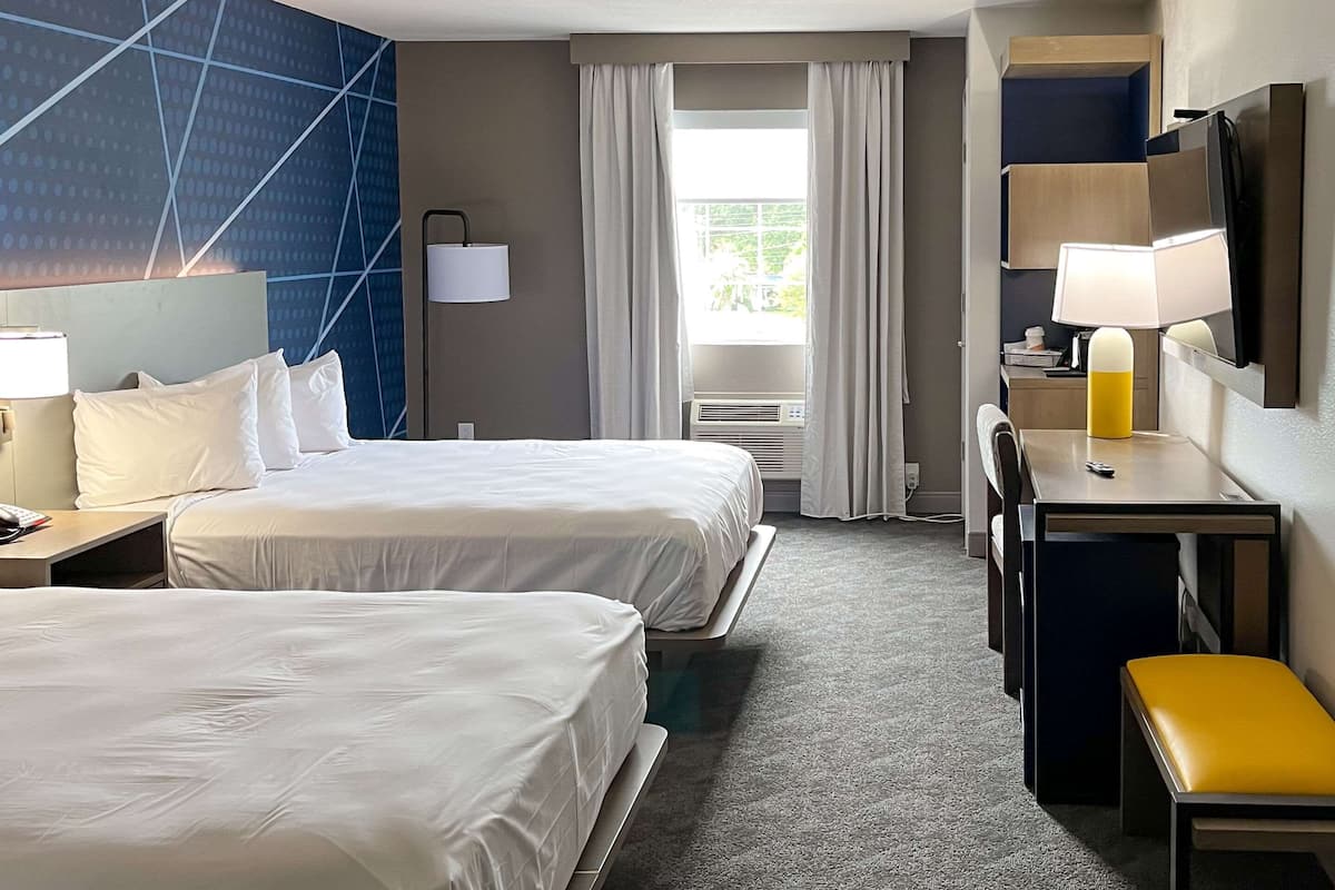 A hotel room with two beds, a desk, a chair, a bench, a TV, a lamp, and a window with curtains.