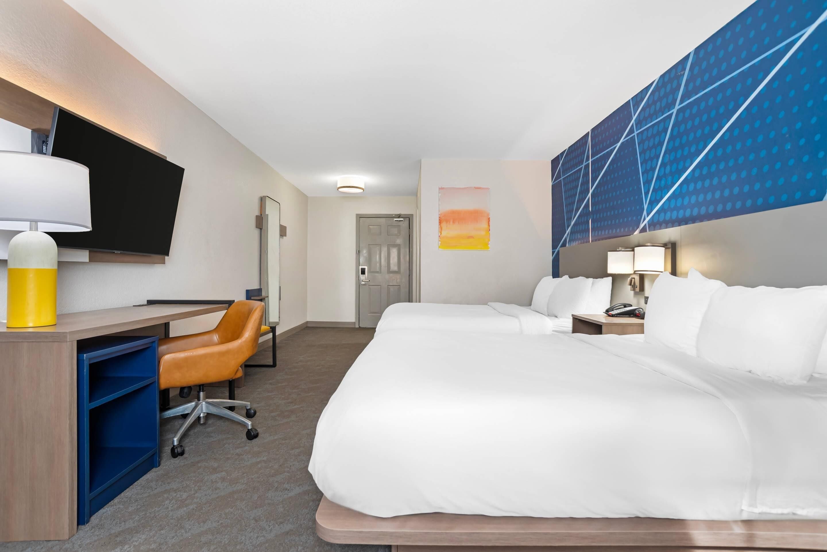 A hotel room with a bed, a desk with a chair, a TV, and a wall with a geometric pattern.