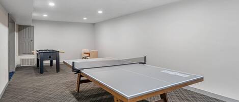 A ping pong table in a room with a carpeted floor, a small table, and a chair.