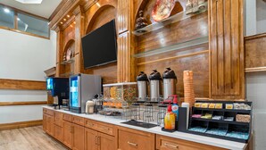 A hotel kitchen with wooden cabinetry, a flat-screen TV, coffee makers, and a beverage dispenser.